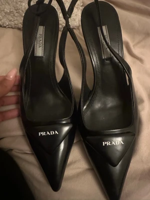 Svarta spetsiga pumps från Prada - Säljer ett par svarta pumps från Prada med spetsig tå och elegant slingback-design. Skorna är i blankt läder och har Pradas logga framtill på tån. Perfekta för dig som gillar lyxiga och trendiga skor med stilren look.