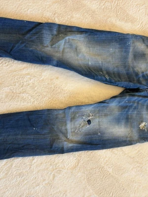Äkta jeans från Dsquared2 - Säljer ett par blå jeans från Dsquared2 med slitningar, hål och färgstänk för en riktigt edgy look. Jeansen har klassisk femficksmodell, knappgylf och cool tvättad finish. Perfekta för dig som gillar streetstyle och statement-plagg.
