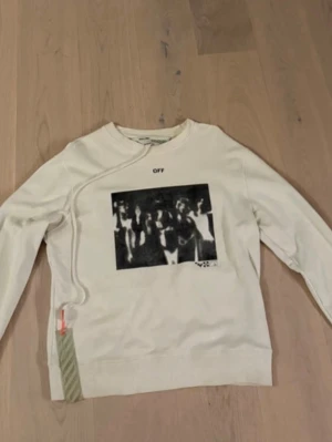 Vit sweatshirt från Off-White - Cool vit sweatshirt från Off-White med svartvit grafiskt tryck på bröstet och texten 'OFF' ovanför. Tröjan har en unik detalj med snedsydd söm och en grön rand med orange tag vid nederkanten. Perfekt för dig som gillar streetwear och statement-plagg.