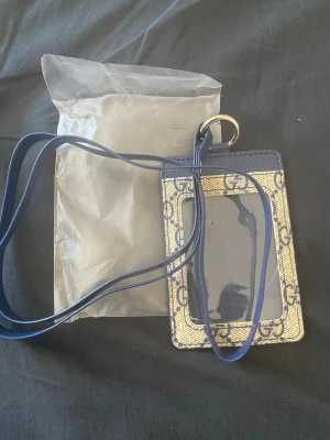 Gucci korthållare med halsband - Snygg korthållare från Gucci med klassiskt GG-mönster i beige och blått. Har ett genomskinligt fack på framsidan och ett blått halsband i läder så du kan bära den runt halsen. Perfekt för kort och ID när du är på språng.