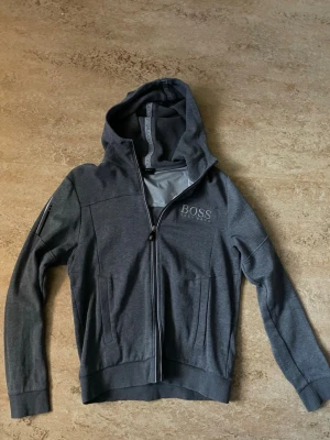 Grå hoodie från BOSS med dragkedja - Grå hoodie från HUGO BOSS i storlek S med dragkedja framtill och två fickor. Märkeslogga på bröstet och huva med fodrad insida. Materialet känns mjukt och bekvämt mot huden. Saknas snören i luvan som syns på bild.