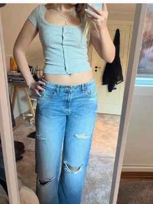 Grå croppad topp med knappar - Snygg grå croppad topp i ribbad material med korta ärmar och knappar framtill. Toppen har en tight passform och en enkel, stilren design som är lätt att matcha med olika outfits. S-M