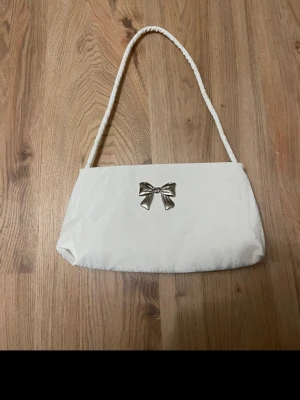 Vit axelväska med rosett - Söt vit handväska med en silverfärgad rosett framtill. Väskan har en avlång, mjuk form och ett matchande axelband. Perfekt för dig som gillar stilrena och gulliga accessoarer.