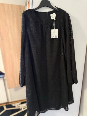 Svart glittrig klänning från UNE - Svart glittrig klänning från UNE med långärmad design och rund halsringning. Klänningen har ett lätt transparent yttertyg med glittereffekt och en rak, lös passform. Perfekt för dig som vill sticka ut med en elegant look.