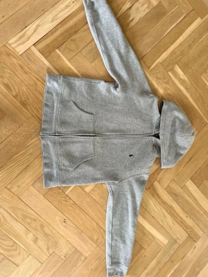 Grå hoodie med dragkedja från Polo Ralph Lauren - Klassisk grå hoodie från Polo Ralph Lauren med dragkedja framtill och två stora fickor. Liten broderad logga på bröstet och mjukt material som känns skönt mot huden. Perfekt för en avslappnad och stilren look.