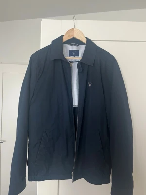 Marinblå vindjacka från Gant - Snygg marinblå vindjacka från Gant med klassisk krage och dragkedja framtill. Jackan har två snedställda fickor och diskret Gant-logga på bröstet. Insidan är fodrad med blå- och vitrandigt tyg. Perfekt för blåsiga dagar.