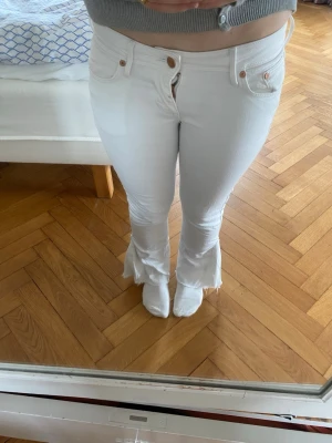 Vita utsvängda jeans med rå kant - Säljer ett par vita jeans med utsvängda ben och råa, fransiga kanter nertill. Jeansen har låg midja, klassiska fickor och knappgylf. De är omsydda från skinny till bootcut. Fråga gärna om bilder om du är osäker