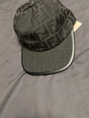 Svart keps från Fendi med logomönster - Snygg svart keps från Fendi med det klassiska FF-logomönstret över hela kepsen. Böjd skärm med diskret kant i läderlook och justerbar rem baktill. Perfekt accessoar för att lyfta din streetwear-stil.