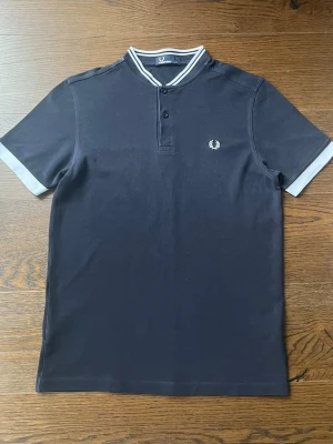 Mörkblå pikétröja från Fred Perry - Snygg mörkblå pikétröja från Fred Perry med korta ärmar och vit logga broderad på bröstet. Tröjan har ribbade muddar och krage med vita detaljer samt två knappar vid halsen. Perfekt för en stilren och sportig look.