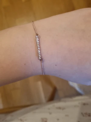 Tunt roséguldigt armband med stenar - Snyggt och minimalistiskt armband i roséguldfärgad metall med en smal rektangulär platta dekorerad med små gnistrande stenar. Justerbar kedja med karbinlås gör att det passar de flesta handleder. Perfekt för dig som gillar stilrena accessoarer.