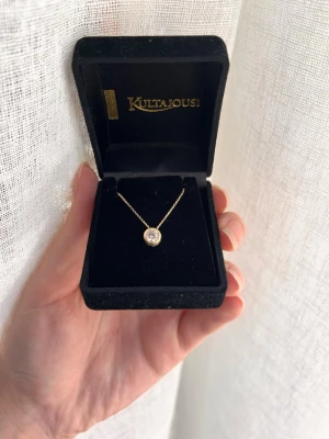 Guldhalsband med klar sten Swarovski - Elegant halsband från swarovski med tunn guldfärgad kedja och en rund klar sten i mitten. Kommer i en svart sammetsask med märkets logga. Perfekt för dig som gillar stilrena och klassiska smycken.
