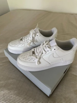 Nike Air Force 1 vita låga sneakers - Helt oaandvända klassiska Nike Air Force 1 sneakers i helvitt läder med perforerad tå, vit sula och matchande vita skosnören. Ikonisk swoosh-logga på sidorna och metall-detalj vid snörningen. Perfekta för en clean och stilren look.   Vart lite för stora för mig. 