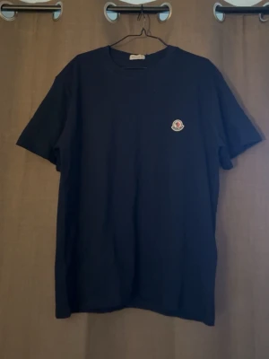 Svart Moncler t-shirt med logga - Säljer en svart t-shirt från Moncler med klassisk rund hals och korta ärmar. På bröstet sitter Monclers ikoniska logga som en snygg detalj. Perfekt basplagg med stilren design och skön passform.