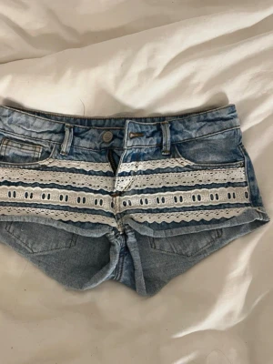 Blå jeansshorts med spetsdetaljer - Snygga ljusblå jeansshorts med vita spets- och broderidetaljer på framsidan. Perfekta för sommaren och ger en cool, bohemisk vibe.