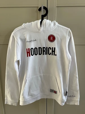 Vit hoodie från Hoodrich med tryck - Snygg vit hoodie från Hoodrich med stor logga i svart och rött framtill samt broderad detalj och patch. Stor ficka på magen och tryck även på ryggen. Klassisk huva och ribbade muddar. Perfekt för en streetwear-look.