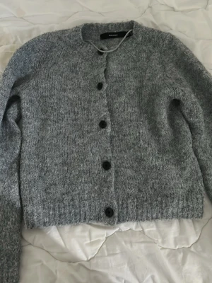 Grå kofta från Vero Moda - Mysig grå kofta från Vero Moda med rund halsringning och svarta knappar framtill. Koftan är stickad i ett mjukt material och har långa ärmar samt ribbade muddar vid ärmslut och nederkant. 