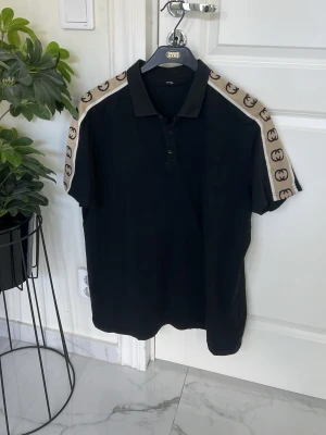 Svart GG pikétröja  - Svart polo t-shirt med krage och knappar framtill. På axlarna och ärmarna finns ett beige band med mörka logomönster som ger en exklusiv känsla. Perfekt för dig som gillar streetwear med lyxig touch.
