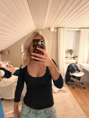 Svart långärmad topp med djup ringning - Snygg svart långärmad topp med djup rund ringning både fram och bak. Figurnära passform som framhäver siluetten. Perfekt att styla med jeans eller kjol för en trendig look.