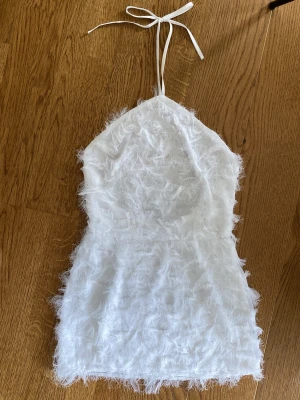 Vit fluffig halterneck kortklänning - Superfin vit halterneck klänning med fluffig textur och knyt i nacken. Klänningen är ärmlös och har en lätt skimrande, fjäderliknande yta som ger en unik look. 