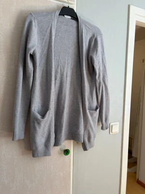 Ljusgrå lång kofta med fickor - Ljusgrå, lång och öppen kofta med två stora fickor framtill. Koftan har ribbade muddar vid ärmslut och nederkant, vilket ger en snygg och avslappnad look. Perfekt att slänga på sig över en t-shirt eller topp.