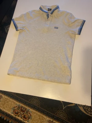 Ljusgrå pikétröja från Hugo Boss - Snygg ljusgrå pikétröja från Hugo Boss med blå detaljer på krage och ärmslut. Tröjan har korta ärmar, knappar vid halsen och broderad logga på bröstet. Perfekt för en stilren och avslappnad look.