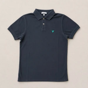 Mörkblå pikétröja från Lyle&Scott - Klassisk mörkblå pikétröja från Lyle n Scott med krage, korta ärmar och knappslå framtill. Tröjan har en liten grön logga broderad på bröstet och är tillverkad i mjuk bomull. Perfekt för en stilren och avslappnad look.