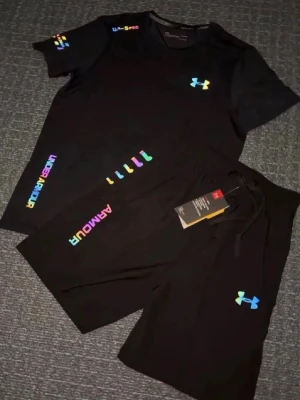 Svart träningsset från Under Armour - Snyggt svart träningsset från Under Armour med färgglada loggor och tryck. Setet består av en t-shirt och shorts, båda med UA-logga i regnbågsfärger och textdetaljer. Perfekt för gymmet eller löprundan. Materialet är lätt och andas.