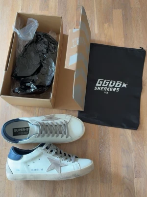 Golden goose superstar - Stilrena vita sneakers från Golden Goose med grå stjärndetalj på sidan, grå skosnören och mörkblå hälkappa. Klassisk låg modell i läder med slitna detaljer och gummisula. Perfekta för en trendig och avslappnad look.