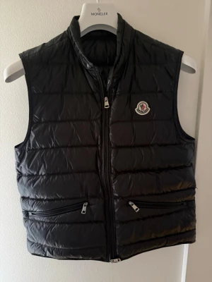 Svart dunväst från Moncler - Snygg svart dunväst från Moncler med klassisk quiltad design och hög krage. Västen har två dragkedjefickor framtill och Monclers ikoniska logotyp på bröstet. Perfekt för lager-på-lager och streetstyle.