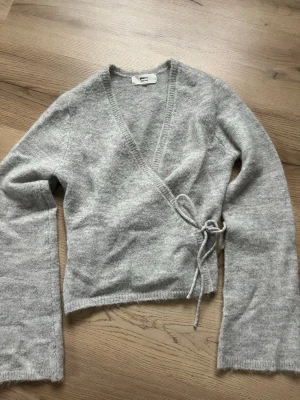 Grå omlottkofta från Gina Tricot - Mysig grå stickad omlottkofta från Gina Tricot med knytband i midjan och v-ringad hals. Koftan har långa ärmar och är tillverkad i ett mjukt, fluffigt material som känns skönt mot huden. Aldrig använd 😊💗