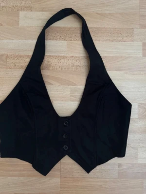 Ny. Svart halterneck väst med knappar - Stilren svart väst i halterneck-modell med djup v-ringning och tre knappar framtill. Västen har en figurnära passform och spetsig nederkant, perfekt för att lyfta din outfit med en edgy touch. Storlek Xl, men den är förminskad som M. Bröst 90-100cm pga gummiband på baksidan.