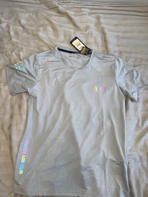 Ljusgrå t-shirt från Under Armour - Snygg ljusgrå t-shirt från Under Armour med holografiska loggor och textdetaljer på bröstet, axeln och sidan. T-shirten har rund halsringning och är kortärmad, perfekt för en sportig look. Materialet är mjukt och känns skönt mot huden. OBS STORLEK L