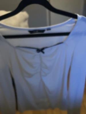 Vit långärmad topp med rosett - Supersöt vit långärmad topp från Gina Tricot med rynkade detaljer framtill och en liten svart rosett vid urringningen. Toppen har en figurnära passform och är perfekt att styla med jeans eller kjol.