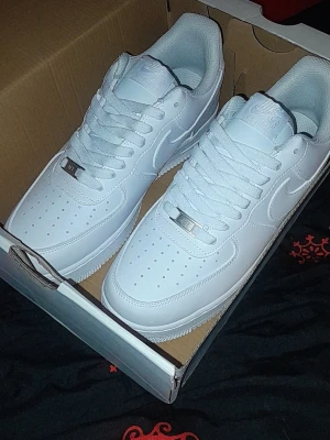 Nike Air Force 1 vita sneakers - Klassiska Nike Air Force 1 sneakers i helvitt läder med perforerad tå, vit sula och matchande vita skosnören. Ikonisk Swoosh-logga på sidorna och metallbricka med 'AF1' på snörningen. Perfekta för en clean och stilren look.