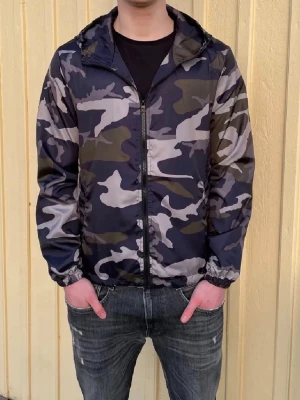 Camo vindjacka med huva - Snygg vindjacka med camouflagemönster i svart, grått och grönt. Jackan har dragkedja framtill, elastiska muddar och en praktisk huva. Perfekt för dig som gillar streetstyle och vill ha en lätt jacka till vardagen.