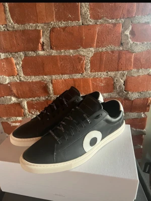 Svarta Oliver Cabell sneakers i läder - Snygga svarta sneakers från Oliver Cabell i äkta läder med vit sula och vit cirkeldetalj på sidan. Skorna har svarta skosnören med logotyptryck och en klassisk låg profil. Perfekta för dig som gillar stilrena och trendiga skor. Har ej använt ofta och de är i helt ny skick. Passar bäst er som gillar grisch/laidback stilen. Nypris: ca3500 VÅRT PRIS🔥: 1600💯