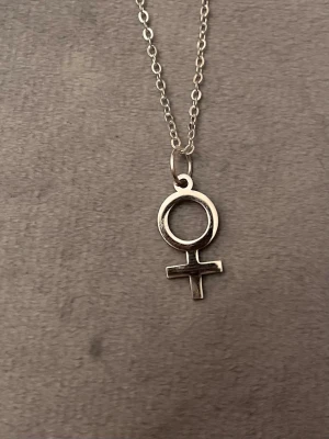 Kvinnosymbolen Halsband Silver imitation Feminist - Fint halsband med kvinnosymbolen. Kedjan är justerbar 40-45 cm och berlocken är ca 1 x 1,8 centimeter. Silverimitation. 10% av det här köpet ger jag till kvinnojouren Karlskrona.  