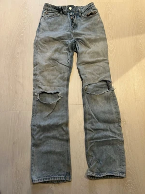 Ljusblåa jeans med hål från H&M - Säljer ett par ljusblåa jeans från H&M med raka ben och stora slitna hål över båda knäna. Jeansen har klassisk femficksdesign och knappgylf. Perfekta för en avslappnad och trendig streetstil.