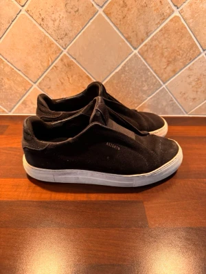 Axel arigato - Säljer mina svarta slip-on sneakers från Axel Arigato i stl 40 med vit sula. Elastisk panel upptill och låg modell som är enkla att ta på och av. Vänster sko har litet hål i bak på sulan som syns på bild 4 , men annars i väldigt bra skick. Original skolåda följer även med.