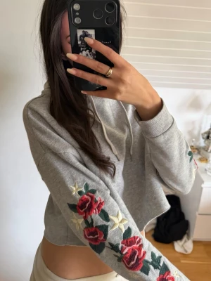 Grå cropped hoodie med rosbrodyr - Grå cropped hoodie med huva och dragsko. Ärmarna har snygga broderade röda rosor och vita stjärnor som ger en unik look. Perfekt för dig som vill sticka ut med detaljer. 