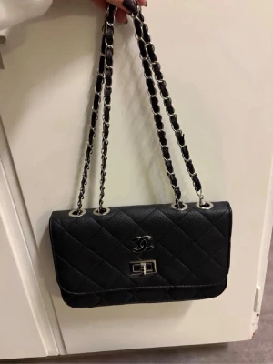 Svårt väska med silverdetaljer  - Helt ny, svart väska med ställbart axelband. Så man kan ha den både som hand-och axelväska. Silverdetaljer. Helt ny, men skada nere i botten på insidan av väskan (se sista bilden). Går säkert att limma om man vill, men inget som syns eller störs, eftersom det är på insidan. Även ett mindre fack på insidan. Inte en skråma i övrigt. Bara stått bland mina andra väskor. Mått ca 25cm lång och ca 16cm hög.