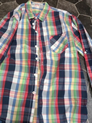 Flerfärgad rutig skjorta stl L - Säljer en färgglad rutig skjorta med lång ärm och klassisk krage. Skjortan har knappar framtill och en bröstficka. Färgerna är blå, röd, grön, gul och vit. Perfekt för dig som vill sticka ut lite extra!