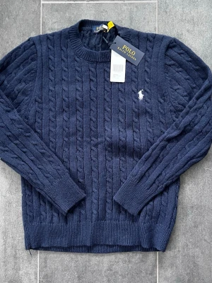 Mörkblå Polo Ralph Lauren - Snygg mörkblå kabelstickad tröja från Polo Ralph Lauren med rund halsringning och klassisk vit logga broderad på bröstet. Tröjan har långa ärmar och ribbade muddar vid ärmslut och nederkant. Perfekt för dig som gillar tidlös och stilren design.