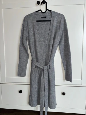 Lång grå kofta LM Sthlm  - Snygg grå lång kofta från Stockh LM med knytskärp i midjan. Koftan har långa ärmar, ribbade muddar och öppen front. 