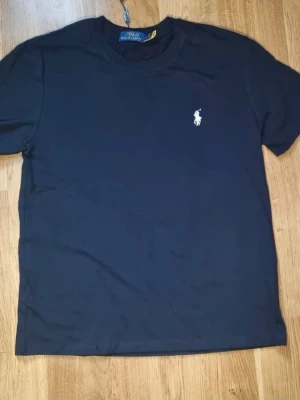 Svart t-shirt från Polo Ralph Lauren - Klassisk svart t-shirt från Polo Ralph Lauren med rund hals och korta ärmar. Diskret logga i vitt broderad på bröstet. Perfekt basplagg med normal passform och mjukt bomullsmaterial.