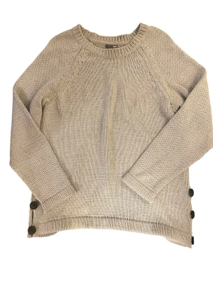 Grå stickad tröja med knappar - Snygg grå stickad tröja med rund halsringning och långa ärmar. Tröjan har dekorativa svarta knappar längs sidorna och ribbade muddar vid ärmslut och nederkant. Perfekt för lager på lager och enkel att matcha till olika outfits.