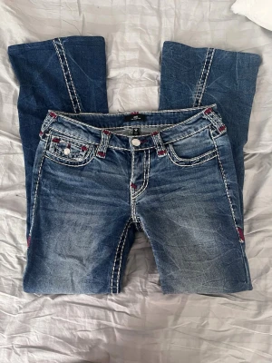 Blå low rise bootcut jeans från True Religion - Säljer ett par blå bootcut jeans från True Religion med tydliga vita kontrastsömmar och röda detaljer vid fickorna.  Jeansen har en jätte fin unik tvätt. Modellen är - Joey, Low Rise Flare! Har även sytt upp dom i längden (se bild) då de var för långa när jag köpte dom, men inget som märks! Skriv vid intresse!💕💕
