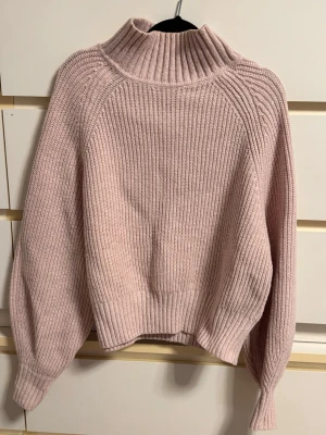 Ljusrosa stickad polotröja - Mysig ljusrosa stickad tröja med hög polokrage och breda ribbade muddar. Tröjan har en avslappnad passform och ballongärmar som ger en trendig look. Perfekt för kalla dagar och enkel att matcha med jeans eller kjol.