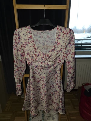Blommig kortklänning från Zara - Supersöt kortklänning från Zara med blommigt mönster i rött, lila och blått på vit botten. Klänningen har v-ringning, långa ärmar och markerad midja som ger en smickrande siluett. Perfekt för vår och sommar! Ena remmen e sönder vid armen, se Bild.