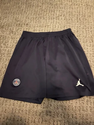 Mörkblå PSG x Jordan shorts - Snygga mörkblå shorts från Jordan i samarbete med PSG. Shortsen har elastisk midja, PSG-logga på vänster ben och vit Jumpman-logga på höger ben. Tillverkade i ett lätt och sportigt syntetmaterial, perfekta för fotboll eller träning.